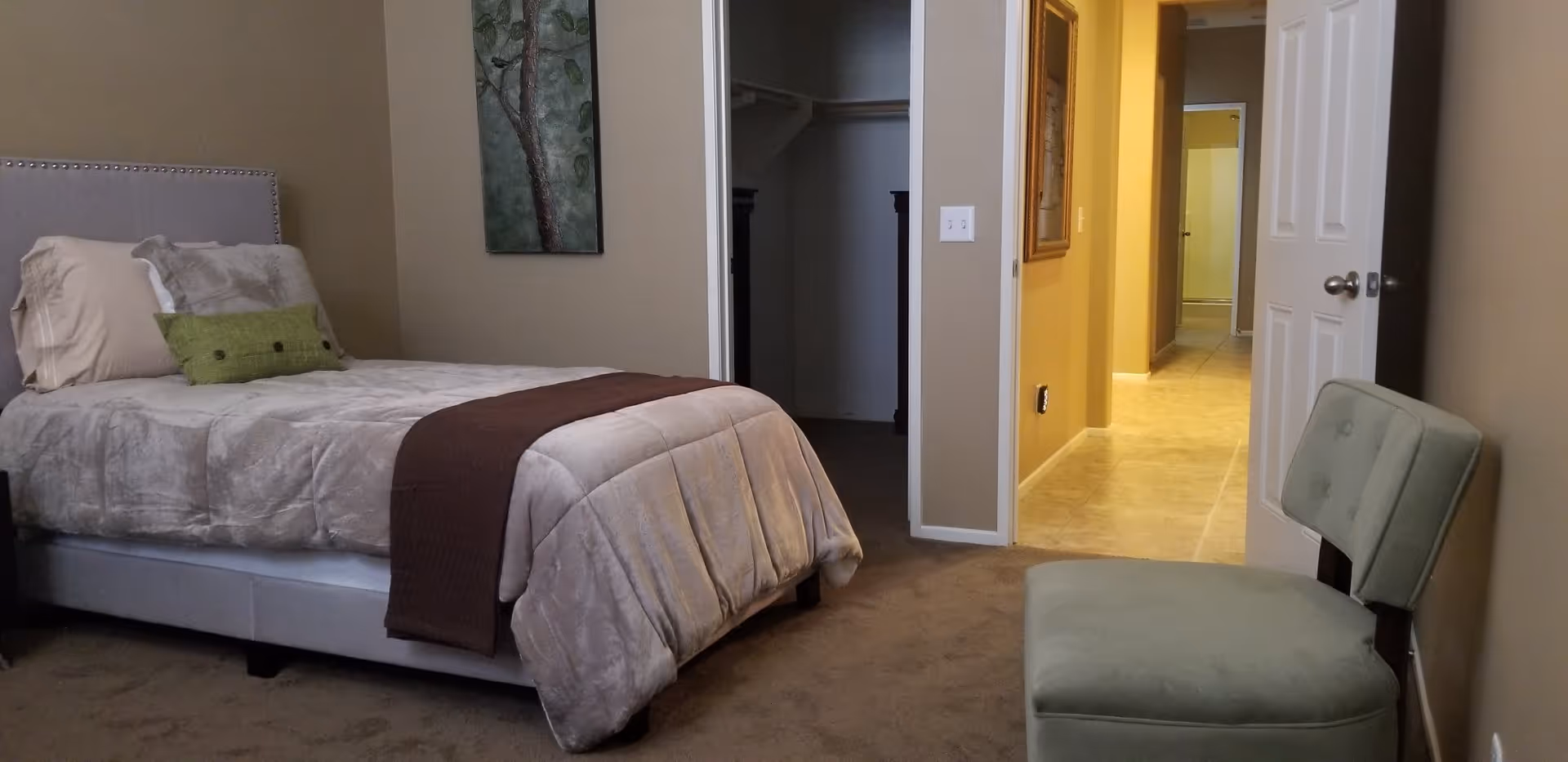 A bedroom with a single bed made with beige and brown bedding, a green decorative pillow, a green cushioned chair, and a painting of a tree on the wall. There is an open door leading to a hallway with tiled flooring and another open door showing a closet.