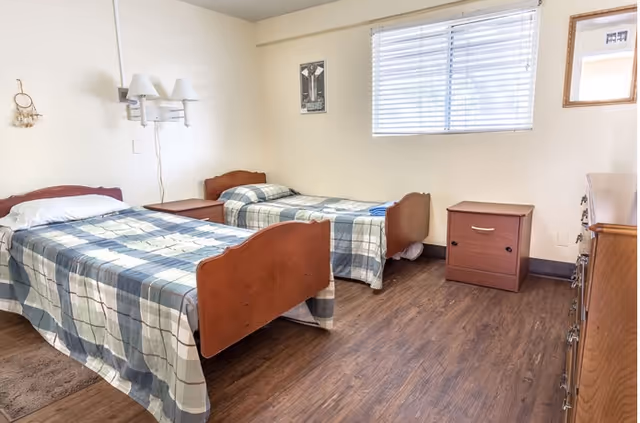 A simple bedroom with two single beds covered in plaid bedspreads, a nightstand between the beds, a window with blinds, a dresser, and a wall-mounted lamp. The floor is wooden and the walls are light-colored.