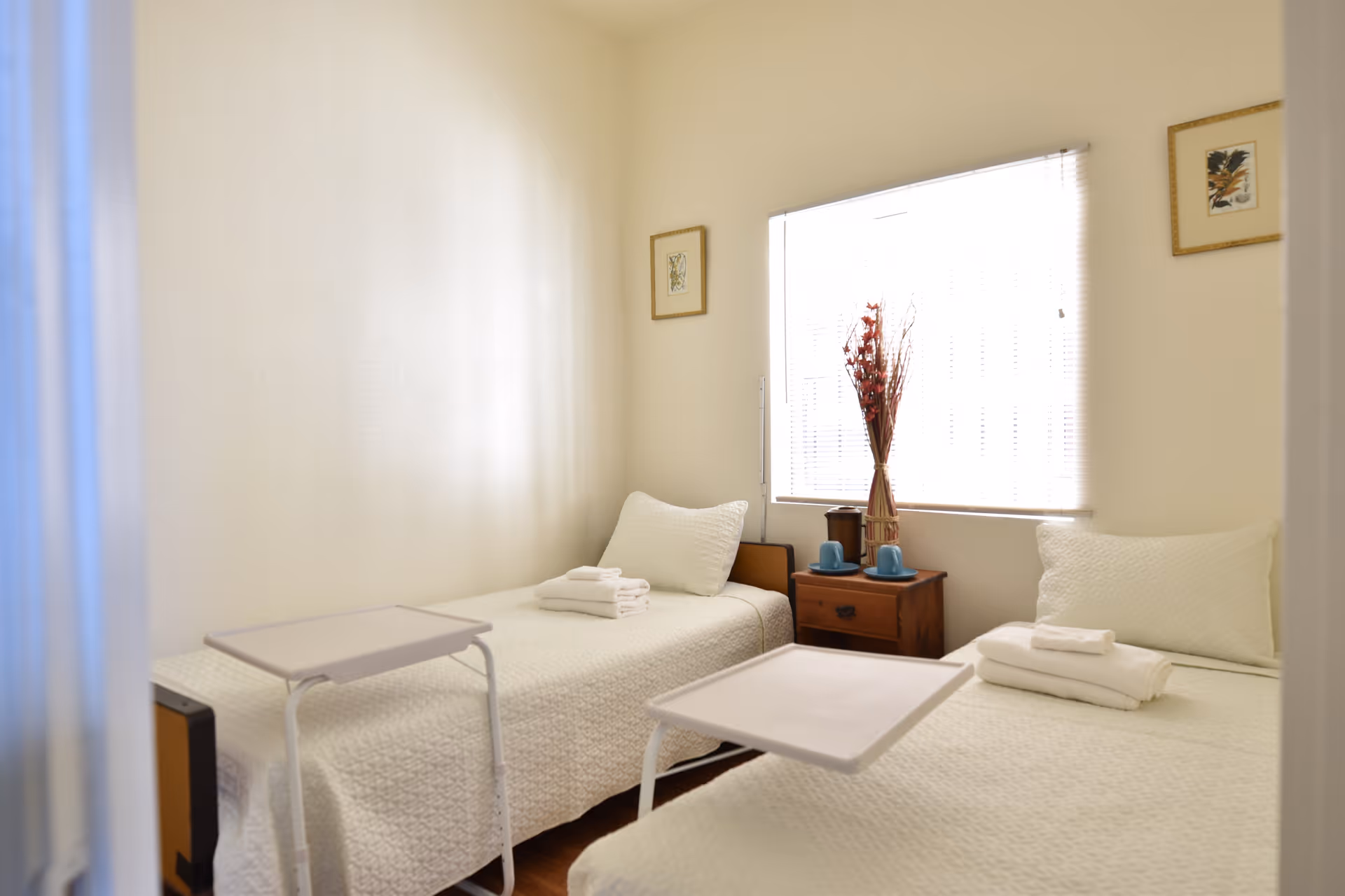 Bright twin bedroom with two single beds dressed in white, a small wooden nightstand under a window, and overbed tables.