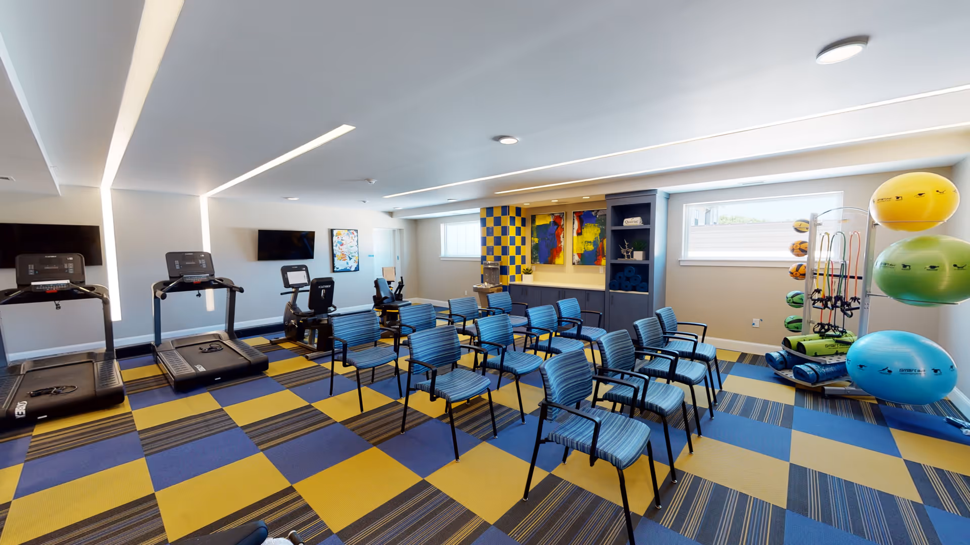 A bright exercise room with blue and yellow checkered carpet, several blue striped chairs arranged in rows, two treadmills, exercise bikes, and a rack holding colorful exercise balls and resistance bands. The room has modern lighting, two windows, and abstract artwork on the walls.