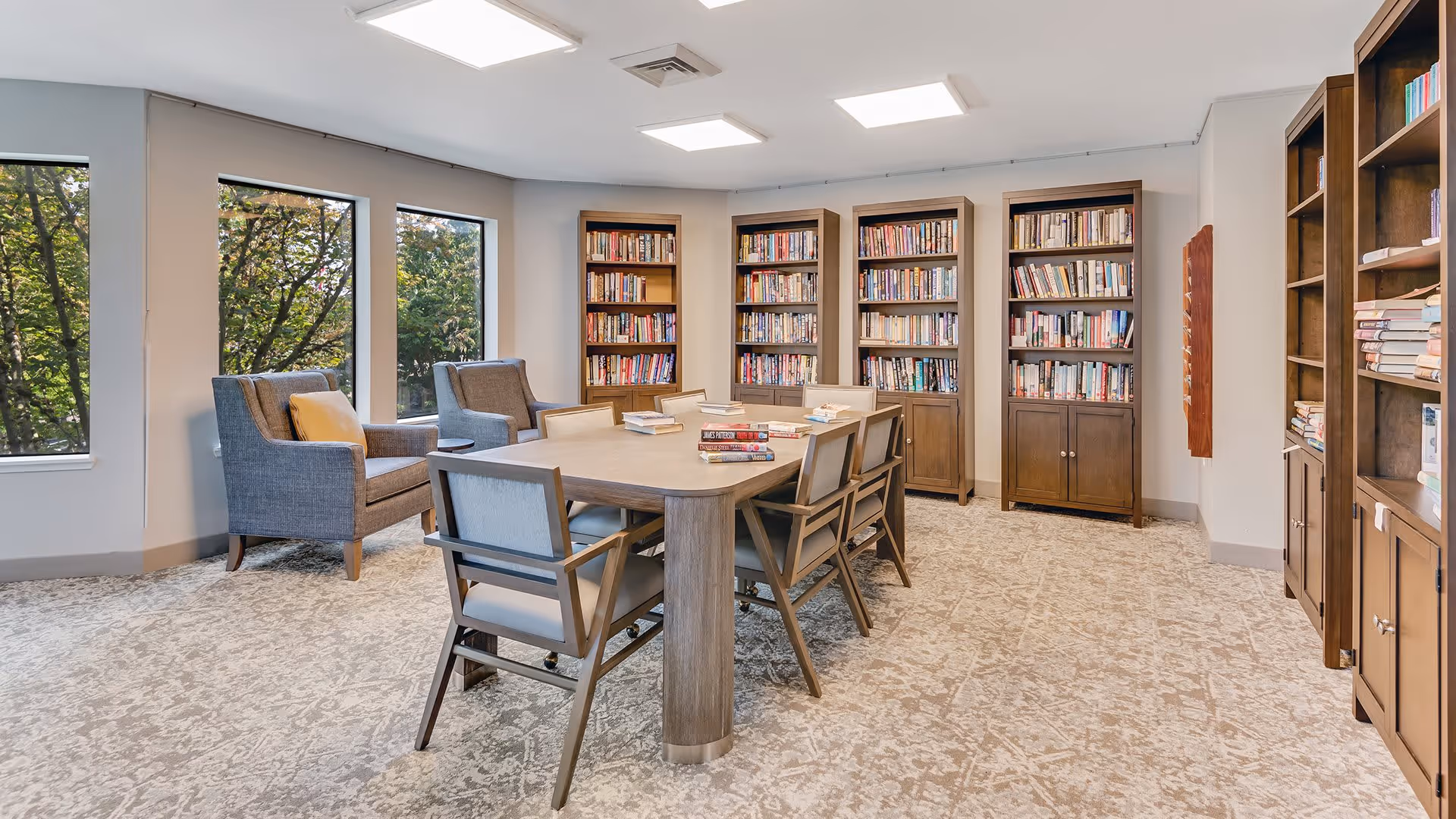 A bright and cozy library room with large windows showing green trees outside. The room features a central wooden table surrounded by six chairs, with several books stacked on the table. Along the walls are multiple tall wooden bookshelves filled with books. Two comfortable armchairs with cushions are placed near the windows.