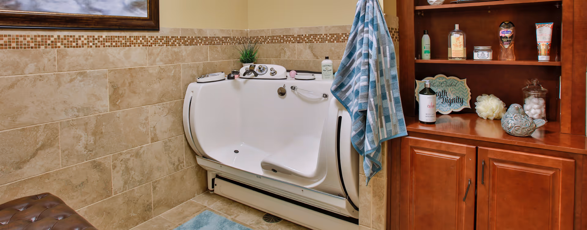 A bathroom with beige tiled walls and floor featuring a white walk-in bathtub with built-in seat and safety handles. A blue and white checkered towel hangs on the side of the tub. To the right, there is a wooden cabinet with shelves holding various toiletries and decorative items. A small blue rug is placed on the floor in front of the tub.