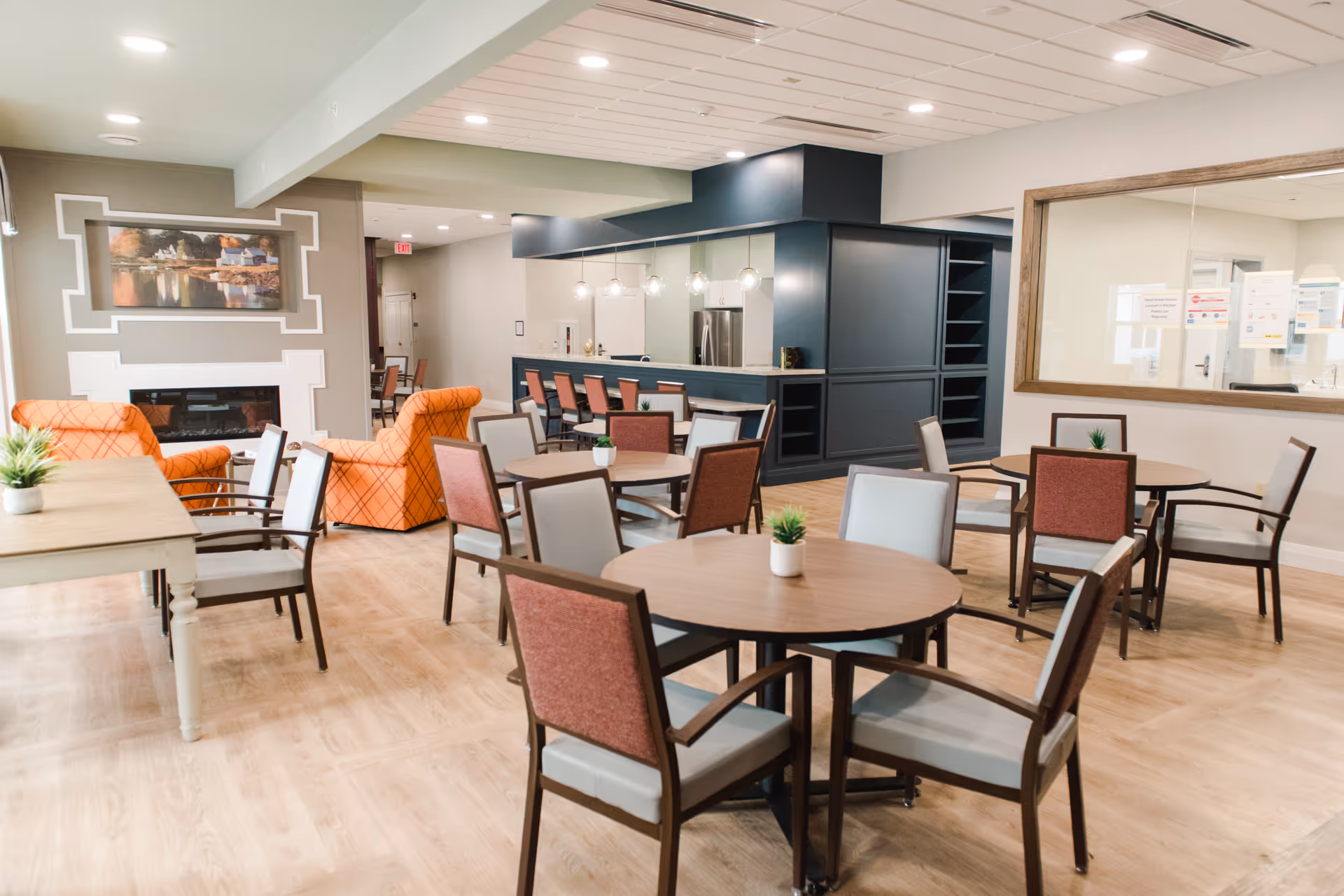 A spacious and well-lit common area in a senior living facility featuring round tables with chairs, small potted plants on each table, a seating area with orange armchairs near a fireplace, and a kitchen area with a counter and bar stools in the background.
