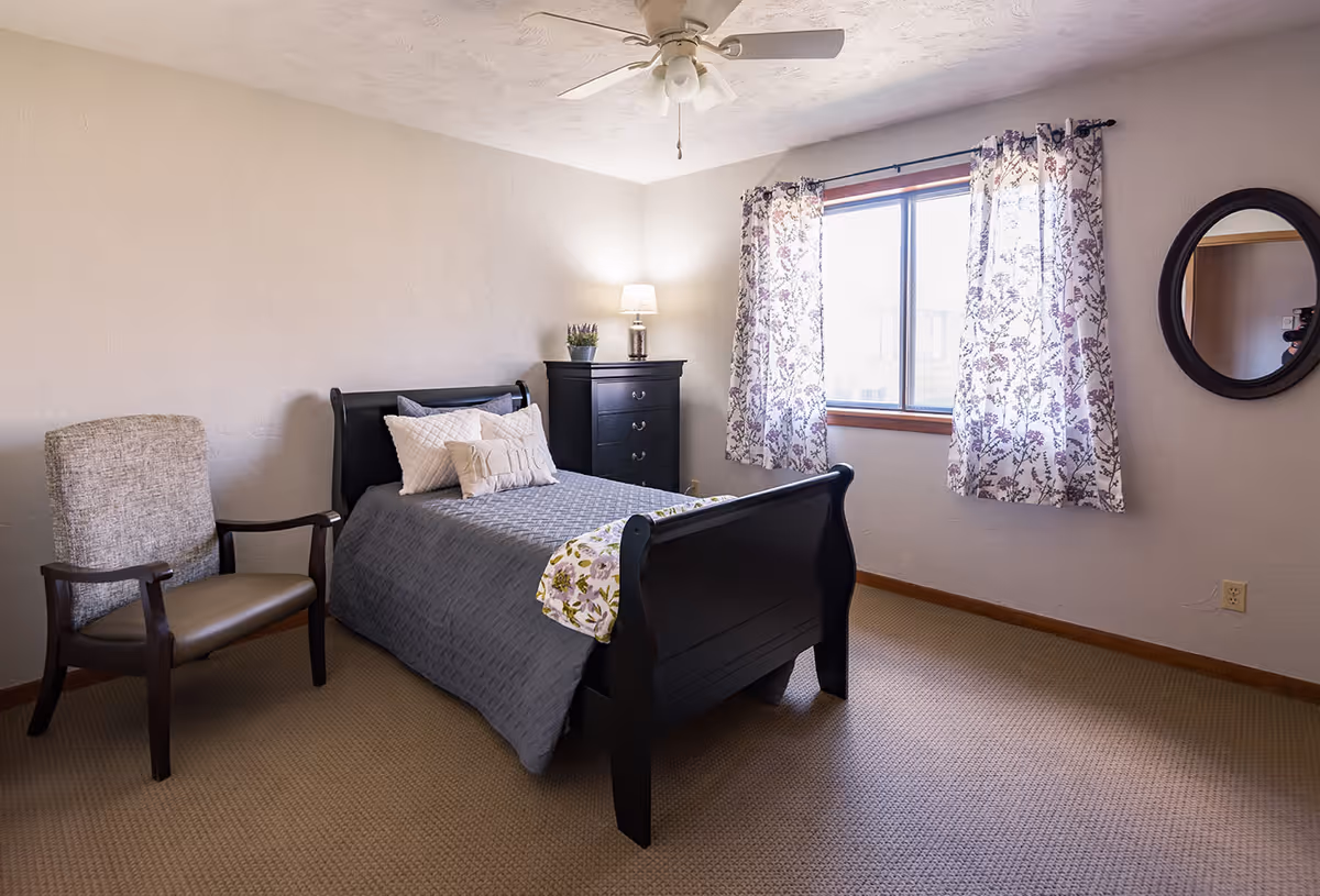A cozy bedroom with a black wooden bed frame, gray bedding, and decorative pillows. There is a cushioned armchair with wooden arms to the left of the bed. A black chest of drawers with a lamp and a small plant sits against the wall near a window with floral curtains. A round mirror hangs on the wall to the right of the window. The room has beige carpet and light-colored walls.