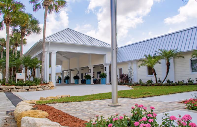Exterior view of West Melbourne Health and Rehabilitation Center showing a covered entrance with white columns, a paved driveway, landscaped garden beds with rocks, palm trees, and flowering plants under a partly cloudy sky.