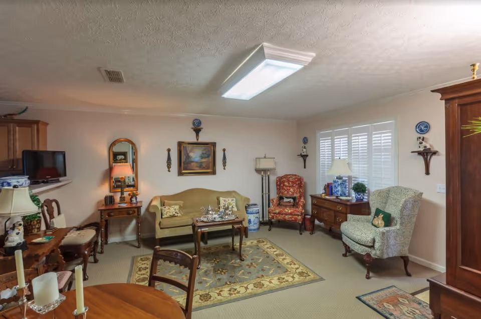 A cozy living room with a beige sofa, two patterned armchairs, a wooden coffee table with a tea set, a wooden side table with a lamp and mirror, a wooden dresser with lamps and decorative items, and a carpet with floral patterns. The room has beige walls, white plantation shutters on the window, and a ceiling light fixture.