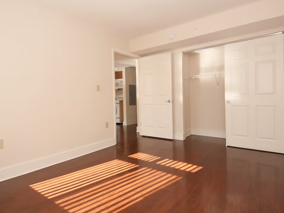 Empty room with wooden floor, white walls, an open closet with sliding doors, and an open doorway leading to a kitchen area.