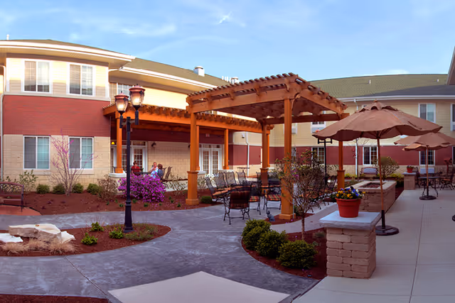 Outdoor courtyard area of a senior living facility with paved walkways, a wooden pergola, patio tables with umbrellas, potted plants, and landscaped bushes and flowers. The building exterior features brick and siding with multiple windows.
