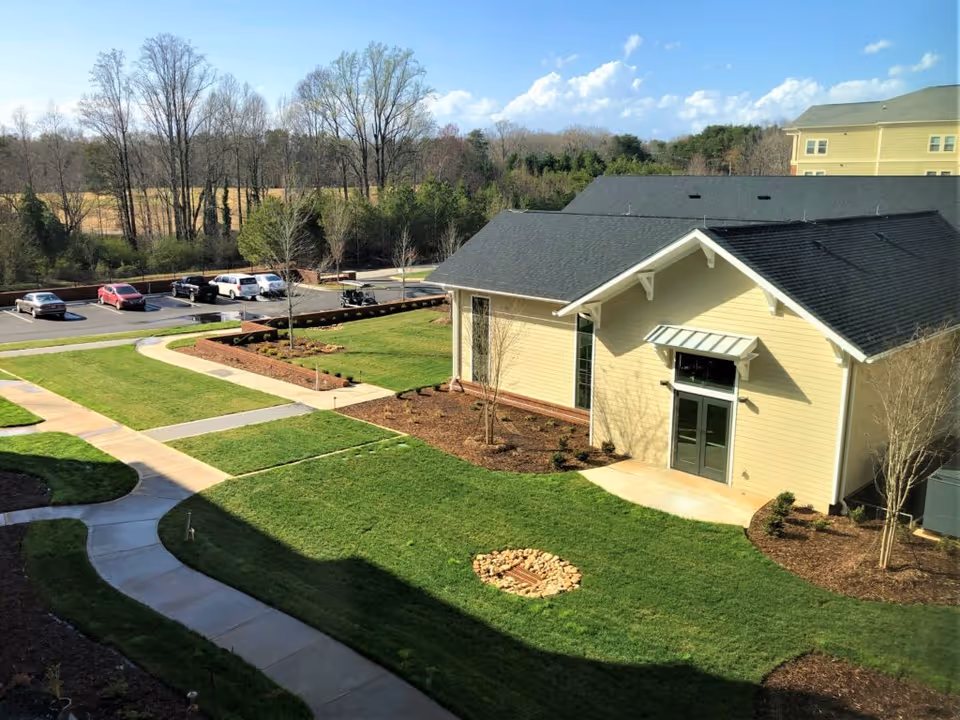 View of a landscaped outdoor area at The Woodlands at Furman, featuring a beige building with a dark roof, green grass, paved walkways, small trees, and a parking lot with several cars in the background under a partly cloudy sky.
