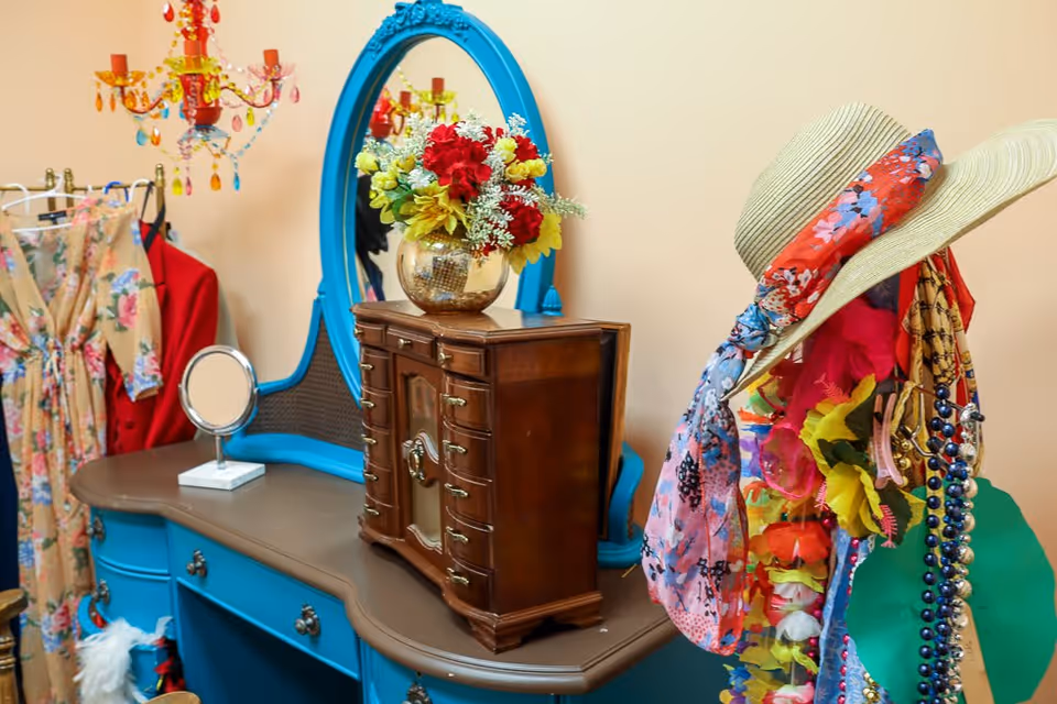A vintage-style blue vanity table with a large oval mirror, a small round mirror, and a wooden jewelry box on top. A colorful bouquet of flowers in a gold vase is placed on the jewelry box. To the right, a hat stand holds a straw hat with a floral scarf and various necklaces and accessories. In the background, a floral dress and a red jacket hang on a clothing rack, and a colorful chandelier is visible above.