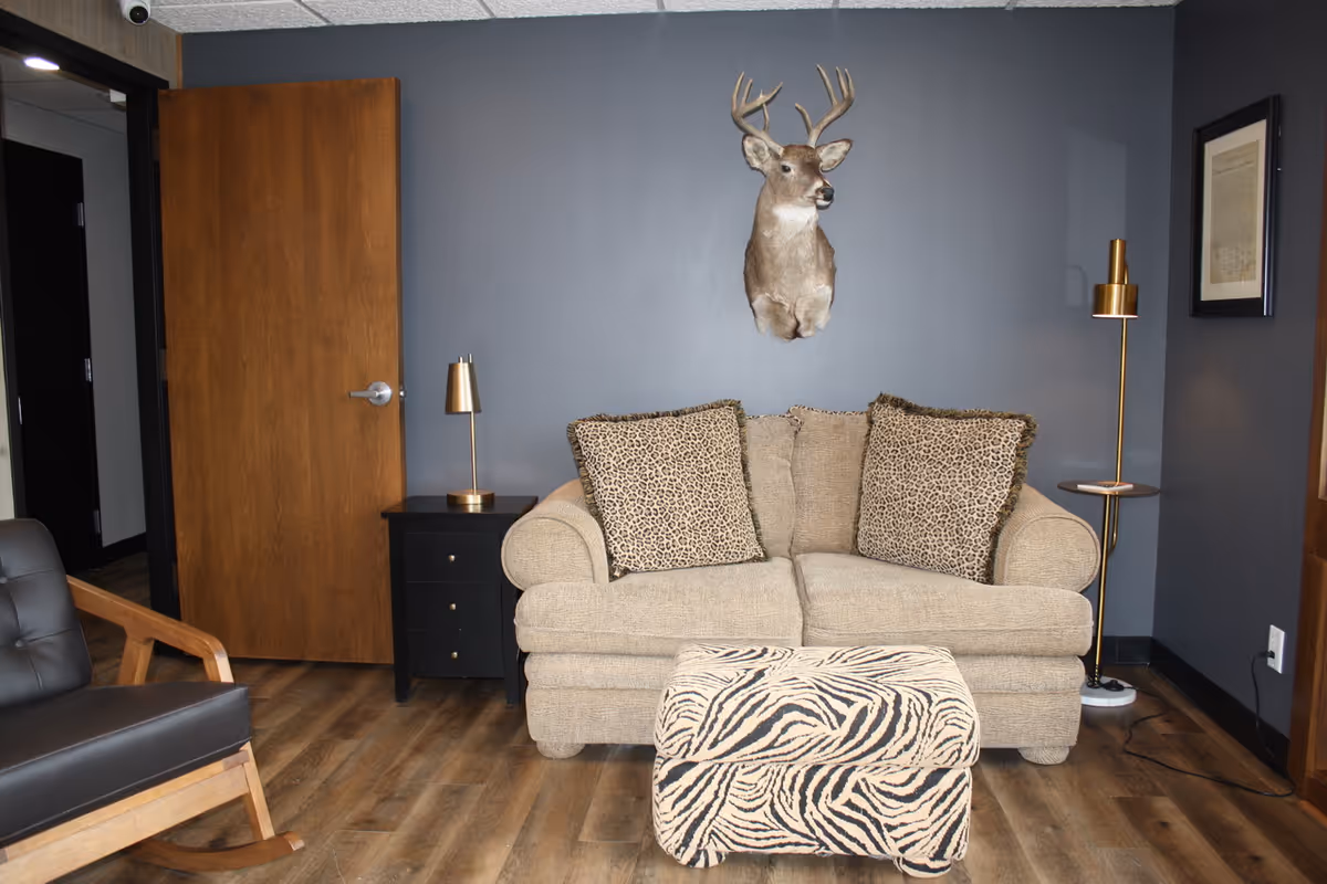 Small sitting area with a beige loveseat, patterned ottoman, animal-print pillows and a mounted deer head on a gray wall.
