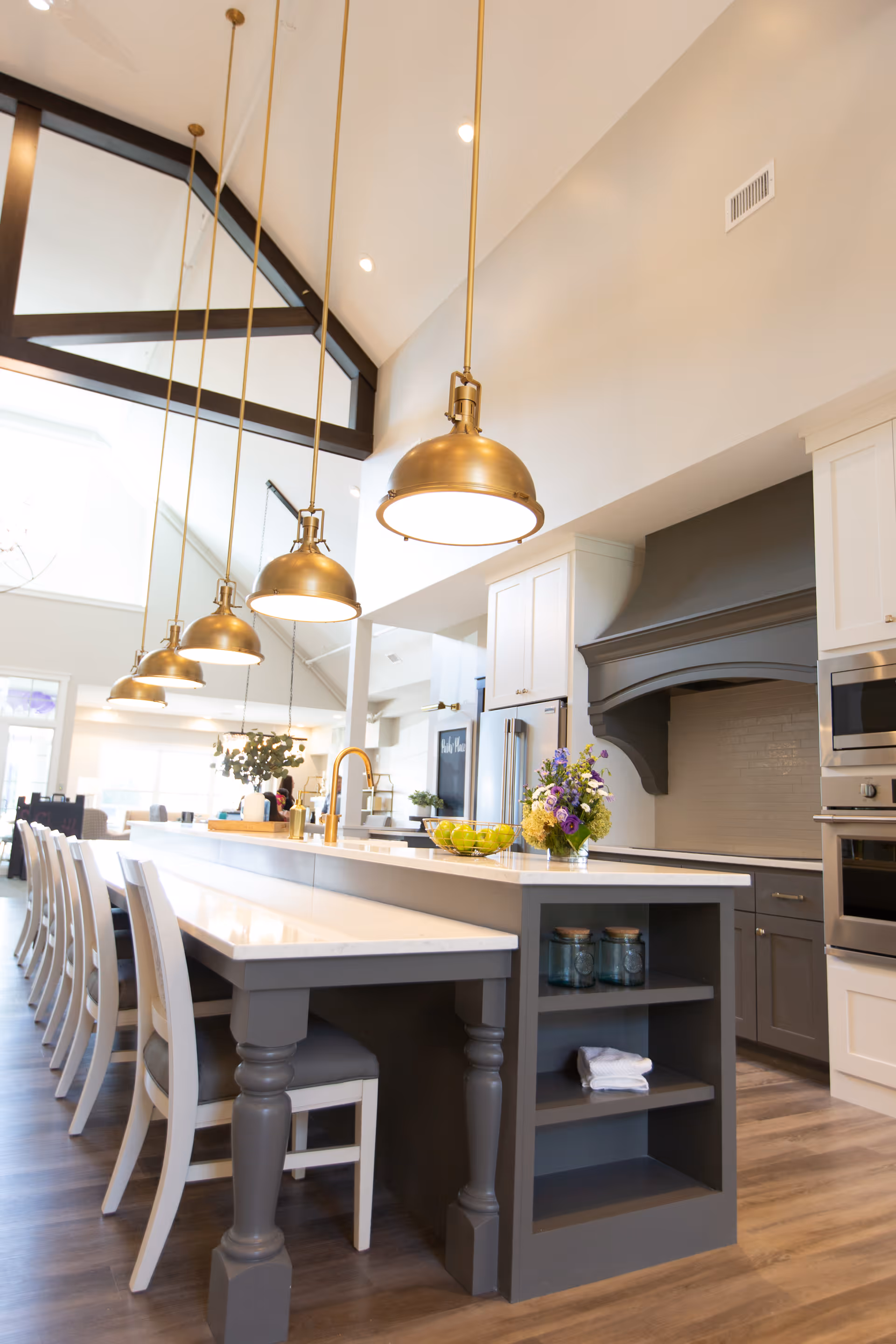 A modern kitchen with a long island countertop featuring white chairs on one side and a built-in shelf on the other. Five brass pendant lights hang from the high ceiling above the island. The kitchen has white and gray cabinetry, stainless steel appliances, a large range hood, and a vase with flowers and a bowl of green apples on the counter.