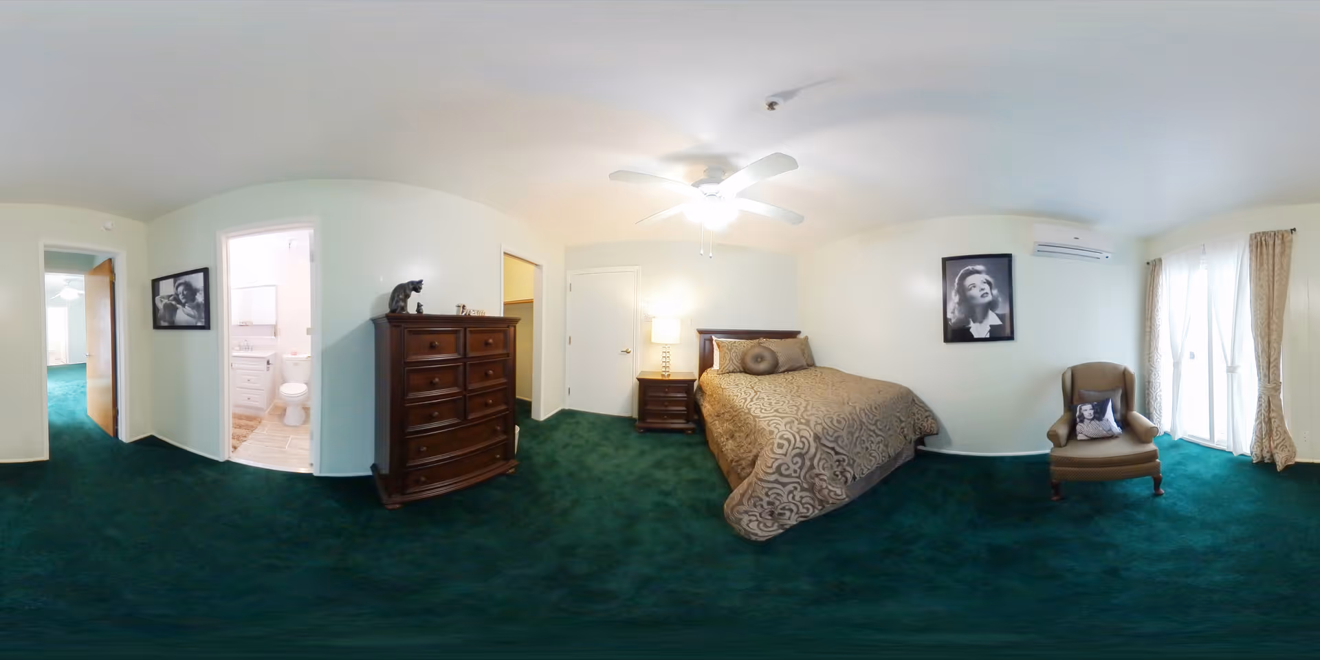 A spacious bedroom with green carpet, a large bed with patterned bedding, a wooden nightstand with a lamp, a wooden dresser with decorative items on top, an armchair with a pillow, and two black and white framed portraits on the walls. There is a ceiling fan with lights, a door leading to a bathroom with a visible toilet, and large windows with curtains allowing natural light into the room.