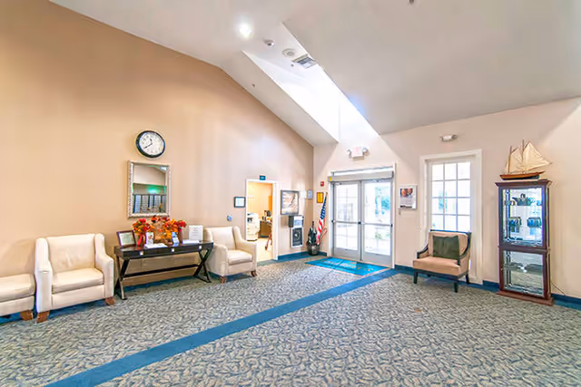 A spacious senior living facility lobby with beige walls and a high ceiling featuring a skylight. The room has a patterned carpet with blue accents, two white armchairs and a black console table with decorative items and flowers. A clock and mirror hang on the wall above the table. There is a glass door entrance with an American flag nearby, a beige armchair with a green cushion, and a glass display cabinet with a model sailboat on top.
