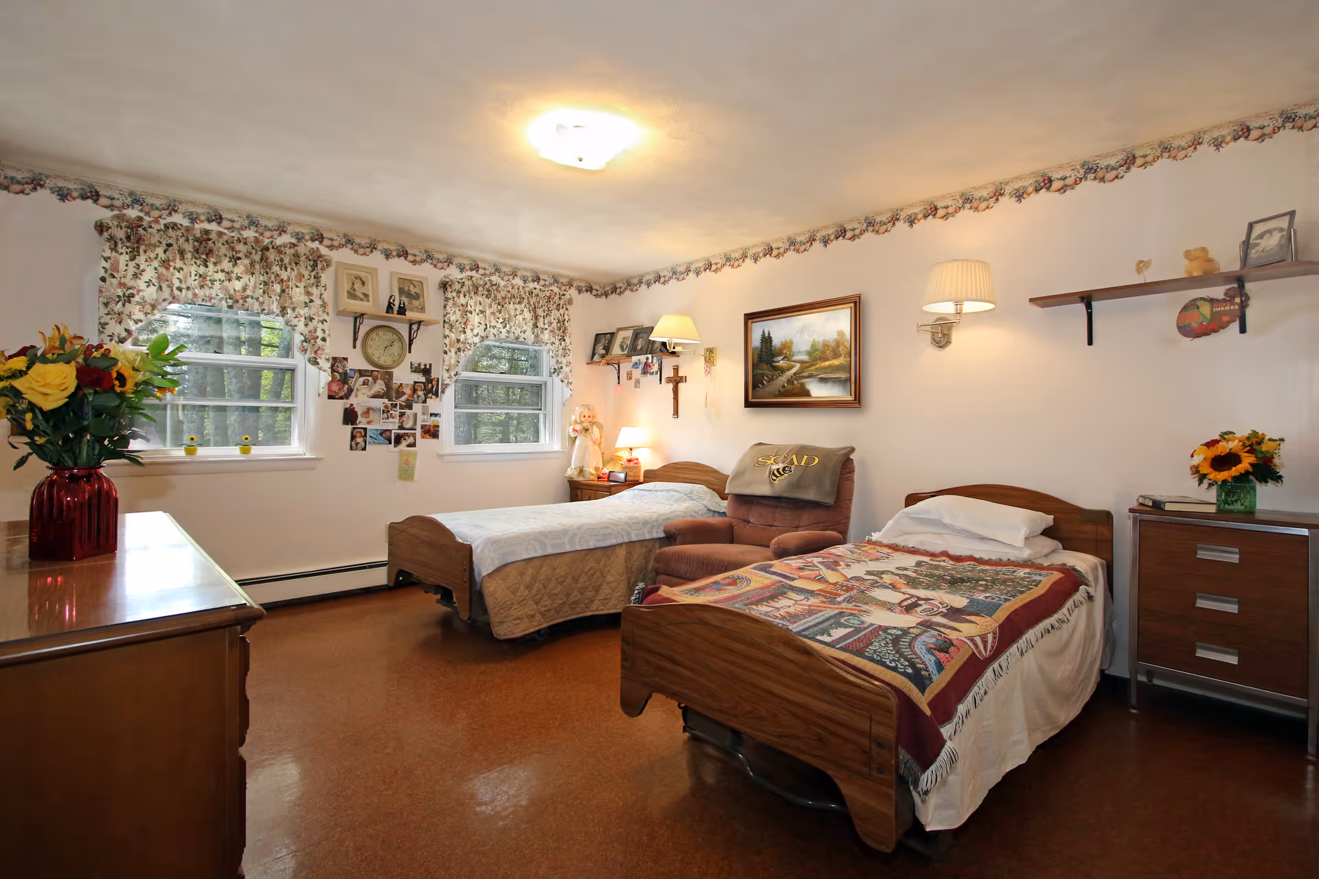 A cozy shared bedroom with two single beds, an armchair, floral curtains, and personal decorations on the walls and dresser.
