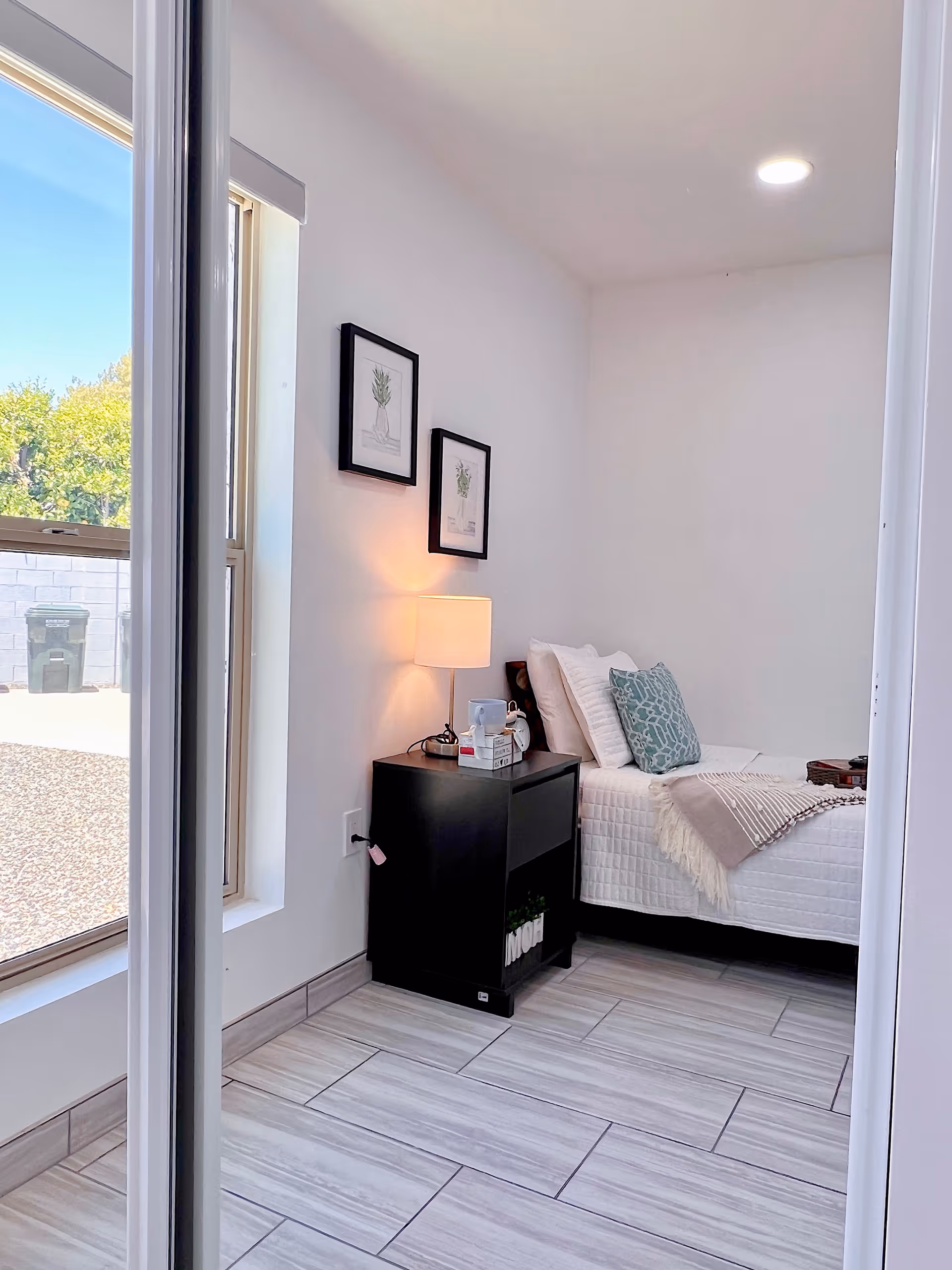 A small, bright bedroom with a single bed against the wall, decorated with white bedding, a teal patterned pillow, and a beige throw blanket. Next to the bed is a black nightstand with a lamp, some books, and a small plant. Two framed botanical prints hang on the white wall above the nightstand. A large window lets in natural light and shows an outdoor view with a trash bin and a white brick wall.
