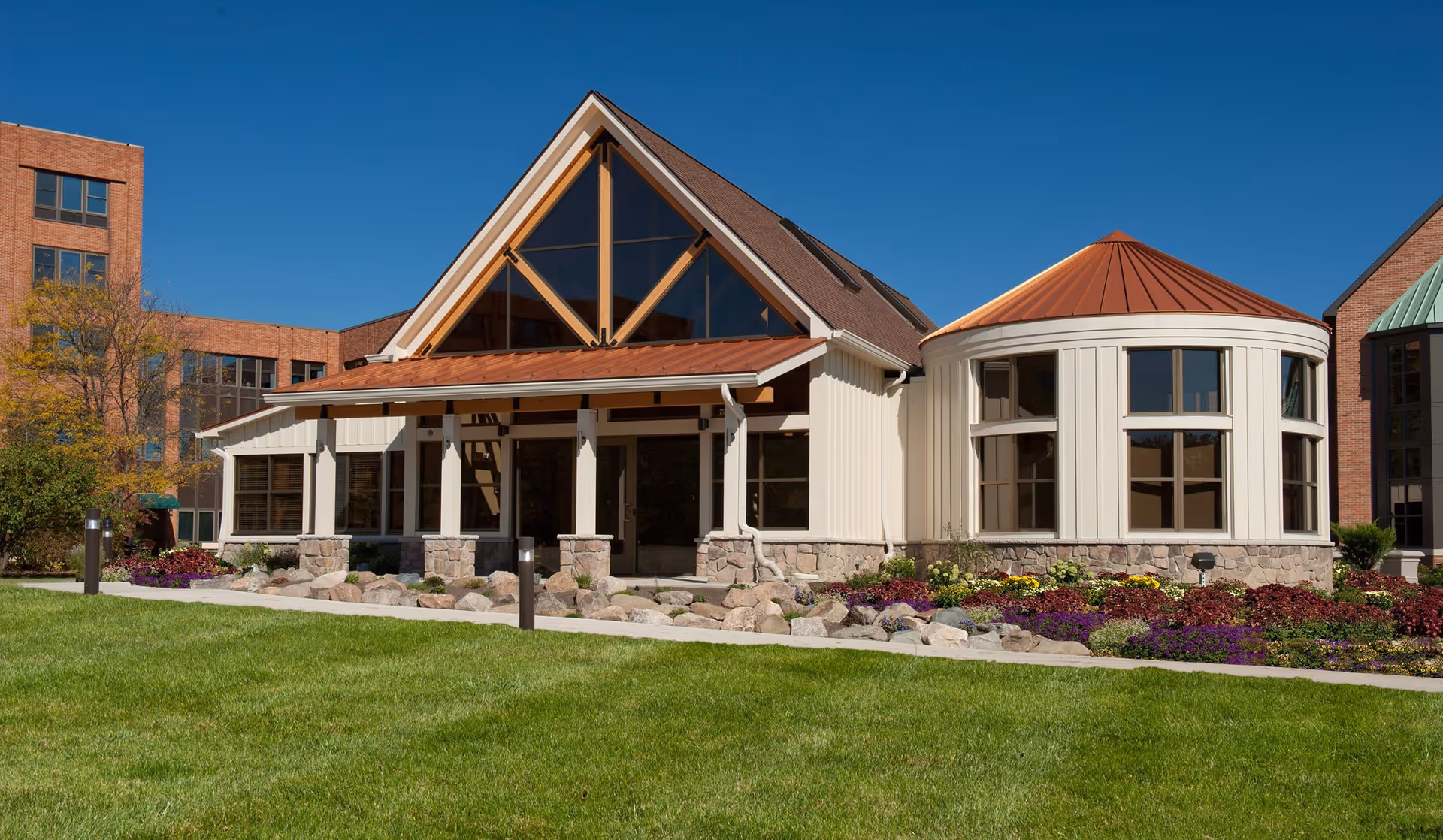 Exterior view of a senior living community building with a triangular roof section and large windows, surrounded by a well-maintained lawn and colorful flower beds under a clear blue sky.