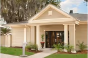 Front entrance of a beige building with white columns, a covered portico, glass double doors, and a landscaped walkway.