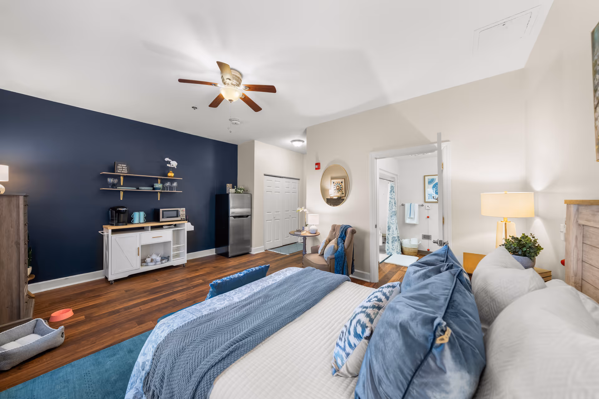 A cozy senior living bedroom with a large bed featuring blue and white bedding and multiple pillows. The room has a dark blue accent wall with shelves holding kitchen items, a small refrigerator, and a microwave. There is a ceiling fan with a light, a comfortable chair with a side table and lamp, and an open door leading to a bathroom with a shower curtain and towels.