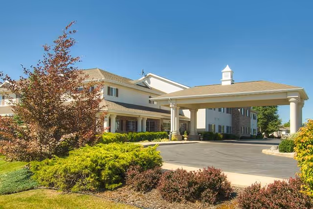 Front exterior of a multi-story senior living building with a covered porte-cochere, landscaped shrubs, and a clear blue sky.