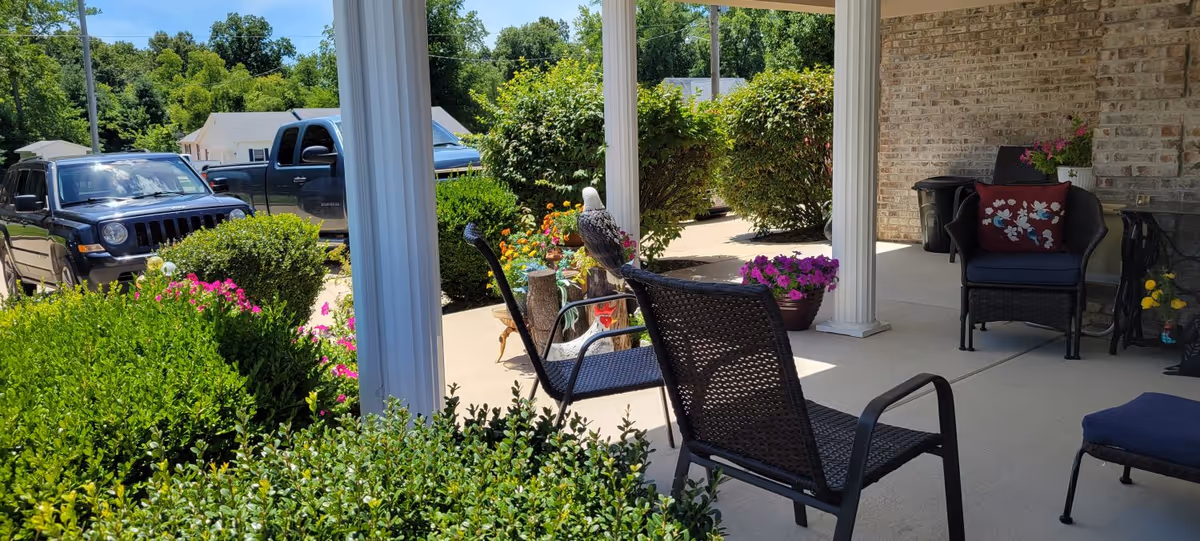 A covered outdoor patio area with black wicker chairs and a small table. The patio is surrounded by green bushes and colorful flowers. In the background, there are parked vehicles and a residential neighborhood visible.