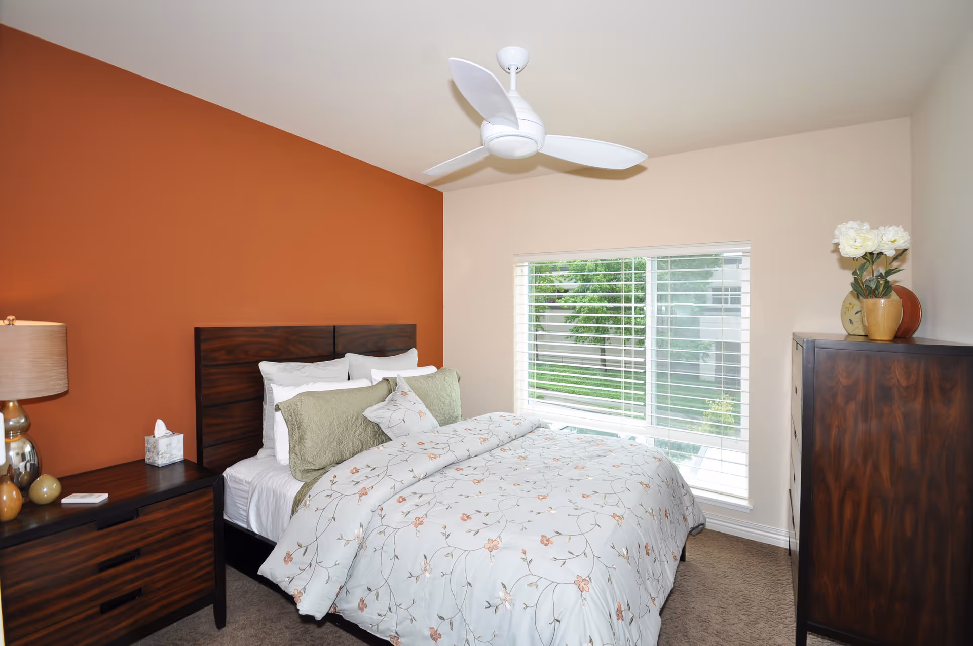 A bedroom with a bed featuring floral bedding and multiple pillows, a wooden headboard, a wooden nightstand with a lamp and tissue box, a wooden dresser with decorative vases and flowers, a large window with white blinds, and a white ceiling fan. One wall is painted orange while the others are beige.
