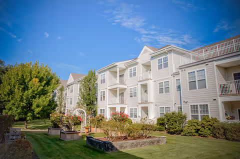 Three-story light-colored residential building with balconies overlooking a landscaped lawn, garden beds, and trees under a blue sky.