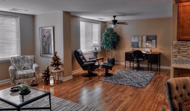 A cozy living room area with wooden flooring, featuring a patterned armchair, a small decorated Christmas tree on a side table, a black leather recliner with matching ottoman, a desk with a chair and decorative items, a large potted plant, and two windows with blinds letting in natural light.