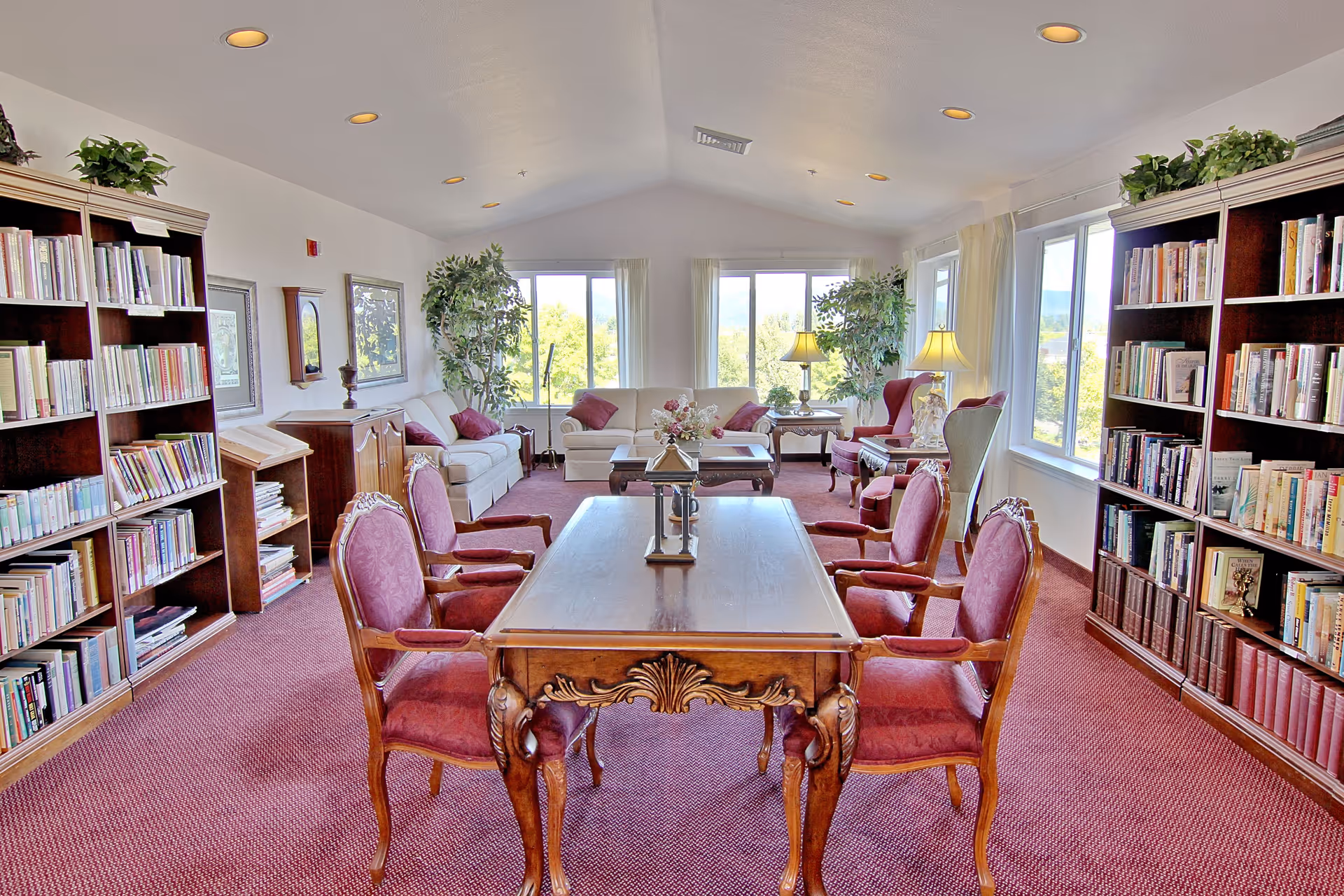 A bright and spacious living room with large windows letting in natural light. The room features a wooden table with four upholstered chairs in the center, two large bookshelves filled with books on either side, and comfortable sofas and armchairs arranged near the windows. There are also potted plants and table lamps adding to the cozy atmosphere.