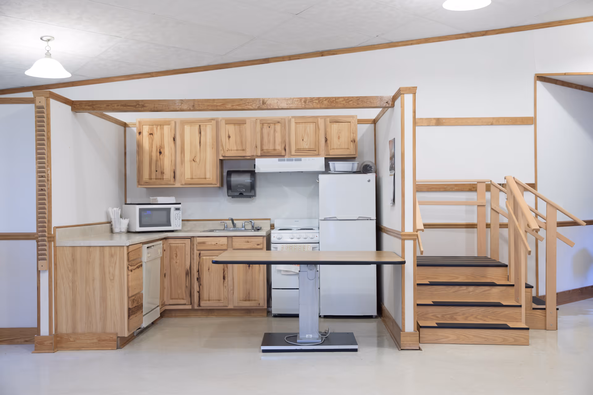 A small kitchen area with wooden cabinets, a white refrigerator, a microwave, a stove with an exhaust hood, a sink, and a dishwasher. There is a table with a metal base in front of the kitchen. To the right, there is a wooden staircase with handrails leading to an upper level. The walls are white with wooden trim.