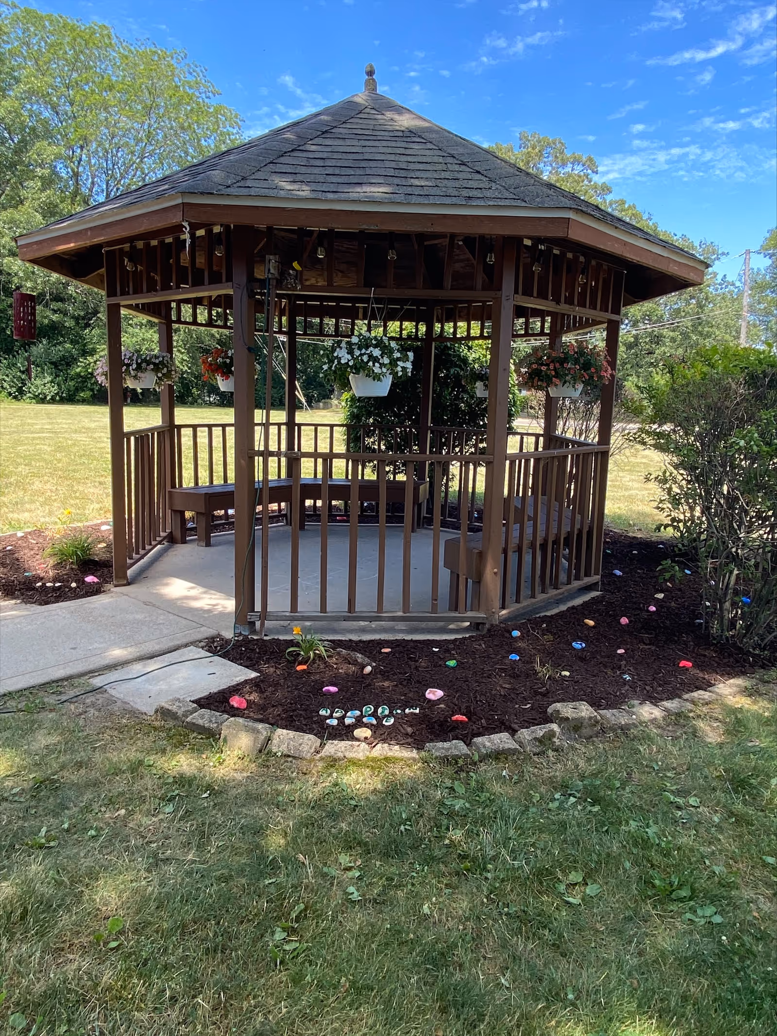 A wooden gazebo with a shingled roof situated in a grassy outdoor area. The gazebo has benches inside and hanging flower pots with white and red flowers. Surrounding the gazebo is a mulched garden bed with colorful painted rocks and small plants. Trees and bushes are visible in the background under a partly cloudy blue sky.