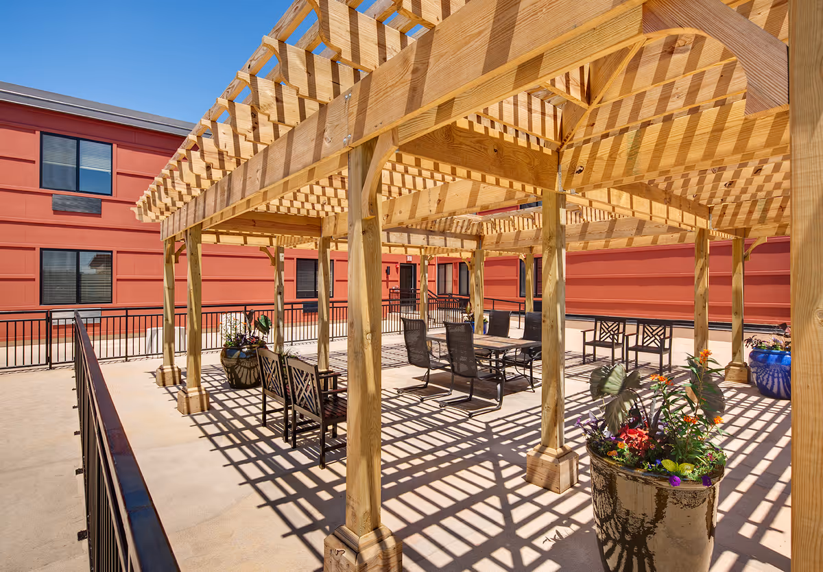 Outdoor patio area with a wooden pergola casting patterned shadows on the ground. The patio has several black metal chairs and tables, along with large planters containing colorful flowers. The background shows a red building with multiple windows.