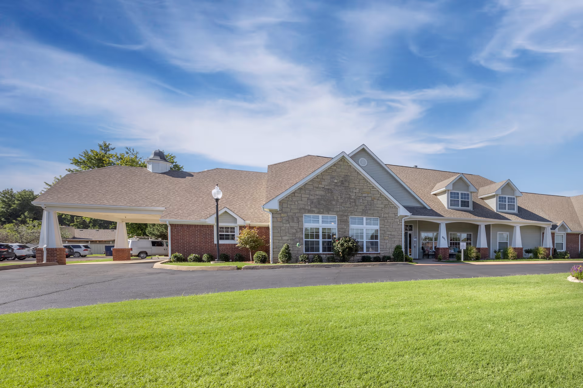 Exterior view of a single-story senior living facility building with a stone and brick facade, a covered entrance, well-maintained green lawn, and a clear blue sky with some clouds.