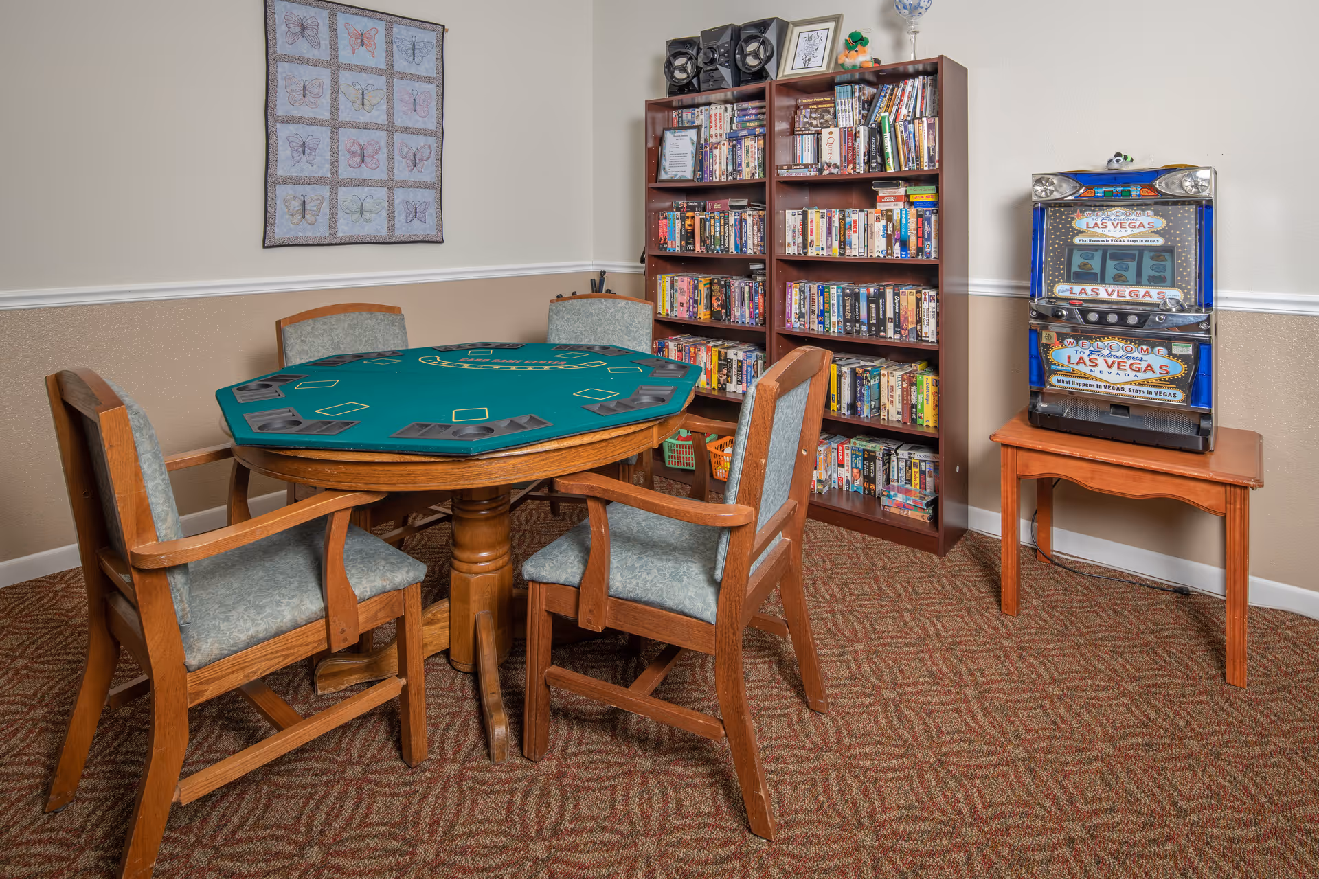 A cozy game room with a round poker table covered in green felt surrounded by four wooden chairs with cushioned seats. Behind the table, there are two tall bookshelves filled with DVDs and books. To the right, a small wooden table holds a Las Vegas-themed slot machine. The walls are beige with a butterfly-themed quilt hanging on one side, and the floor is carpeted with a patterned design.