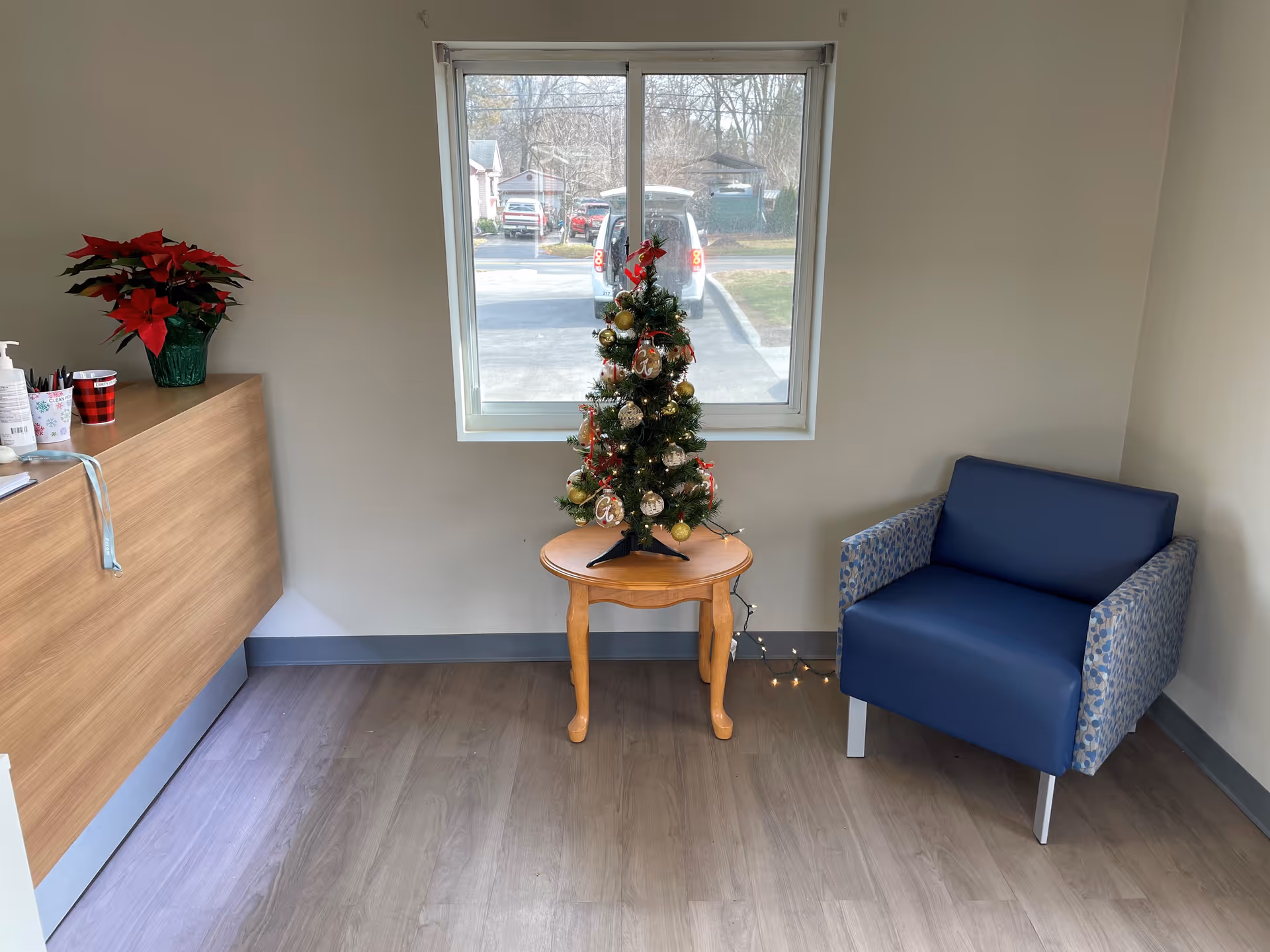 A small room with a window showing a parking lot outside. Inside, there is a wooden table with a decorated small Christmas tree on it, a blue armchair with patterned armrests to the right, and a wooden counter with a poinsettia plant and some cups on the left.