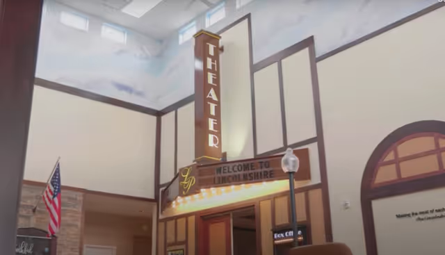 Interior view of a senior living facility featuring a theater entrance with a vertical sign reading 'THEATER' and a marquee sign that says 'WELCOME TO LINCOLNSHIRE'. The walls are decorated with a sky mural near the ceiling, and an American flag is visible on the left side.