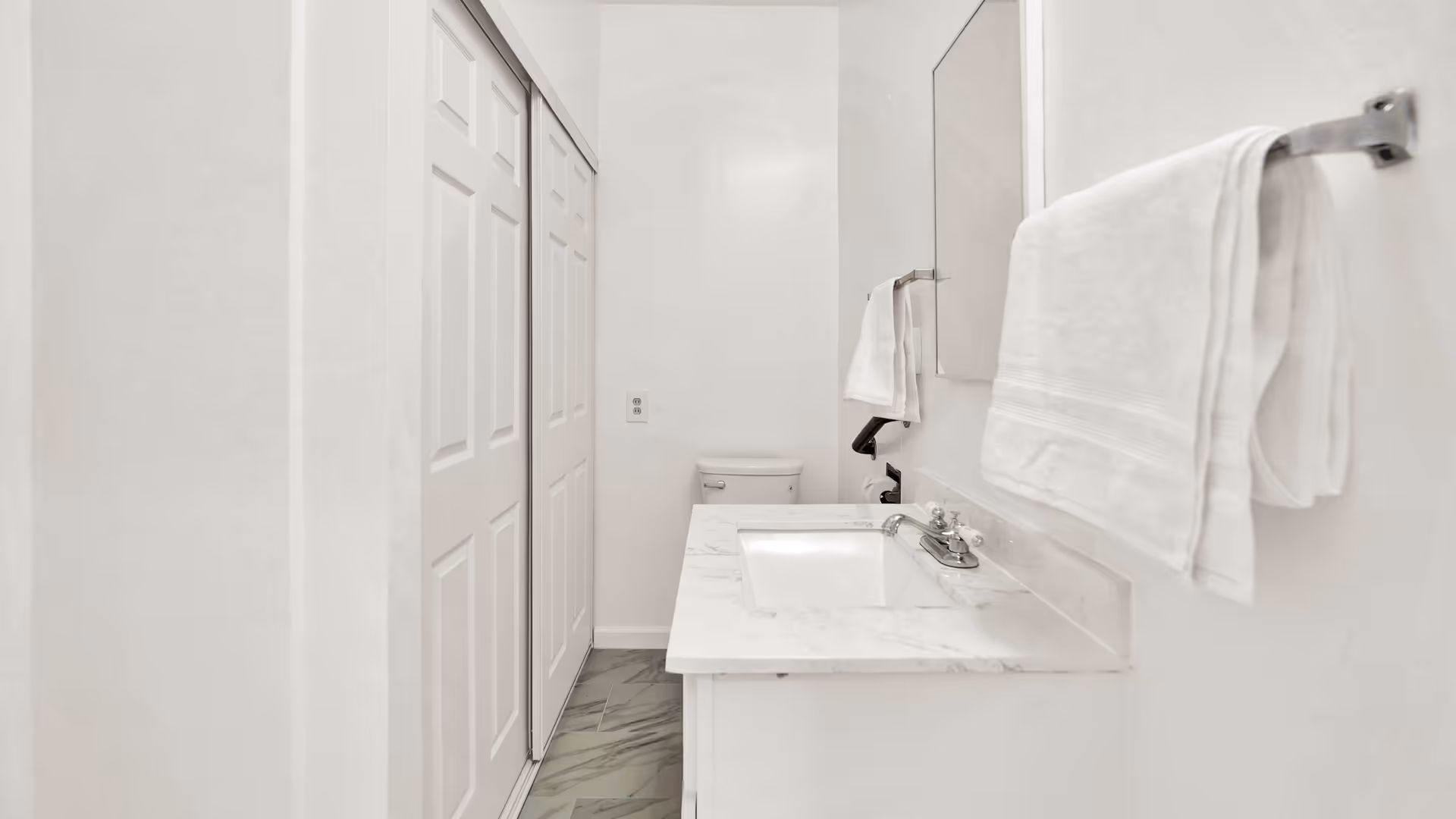 A clean, white bathroom featuring a marble countertop with a built-in sink, a large mirror above the sink, a towel rack with a folded white towel, a toilet in the background, and sliding closet doors on the left side.