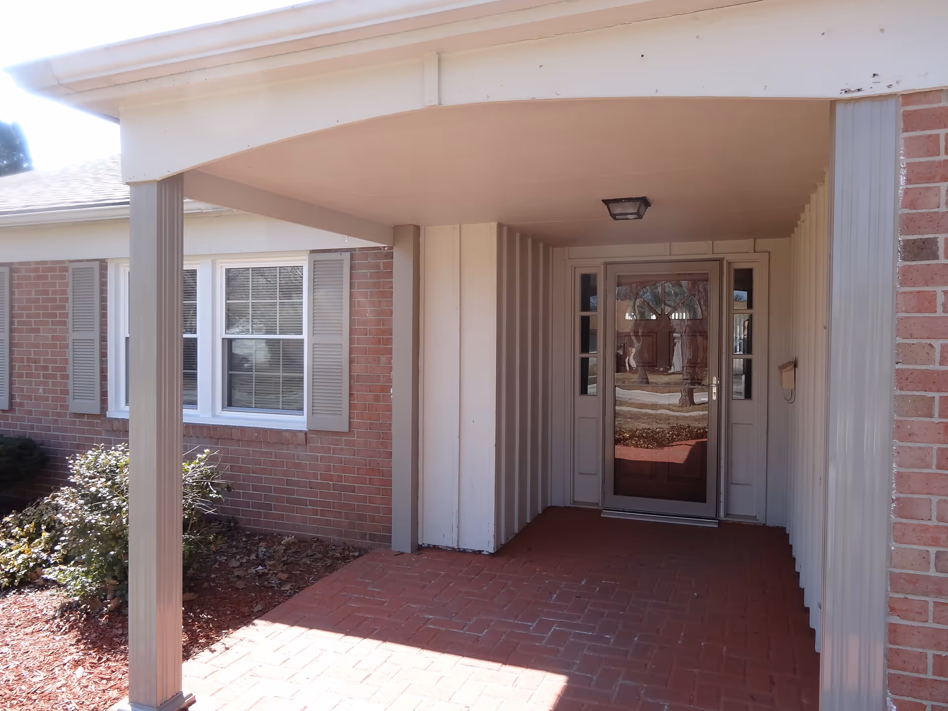 Covered entrance area of a brick building with a glass front door and sidelights, a small porch light on the ceiling, and a window with shutters to the left. There are some bushes and mulch along the side of the building.
