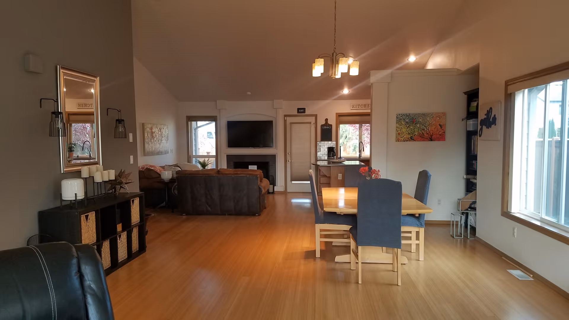Interior view of a senior living facility showing a combined living and dining area with wooden flooring. The living area has brown sofas facing a wall-mounted TV above a fireplace. The dining area features a wooden table with four chairs covered in blue fabric. There is a kitchen visible in the background with a window and a door labeled 'EXIT'. The walls are decorated with paintings and a mirror, and there is a large window on the right side letting in natural light.
