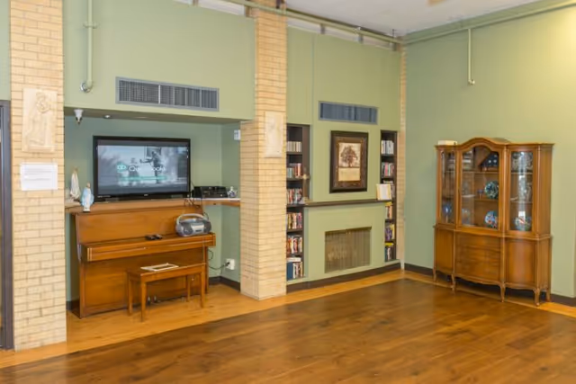 A common living area with a piano beneath a wall-mounted TV, built-in shelves around a fireplace, a china cabinet, and hardwood floors.