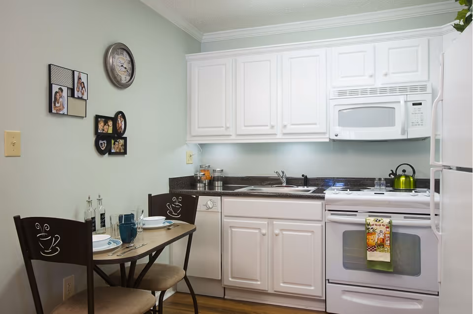 A small kitchen area with white cabinets, a white stove with a green kettle on top, a white microwave above the stove, and a white refrigerator. There is a small dining table with two chairs, each with a coffee cup design on the backrest. The table is set with plates, glasses, and utensils. On the wall above the table are several framed family photos and a round clock.