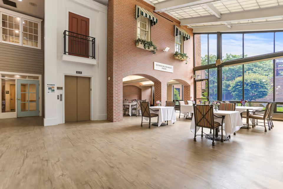 Bright dining area with several tables covered in white tablecloths and chairs arranged around them. The room features large floor-to-ceiling windows letting in natural light and offering a view of greenery outside. There is a brick wall with two windows and flower boxes, and an elevator with closed doors on the left side of the image.