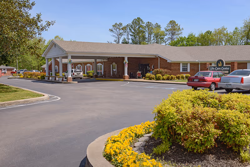 Exterior view of the Life Care Center of Centerville building with a covered entrance, several parked cars, landscaped flower beds with yellow flowers and green shrubs, and trees in the background under a clear sky.