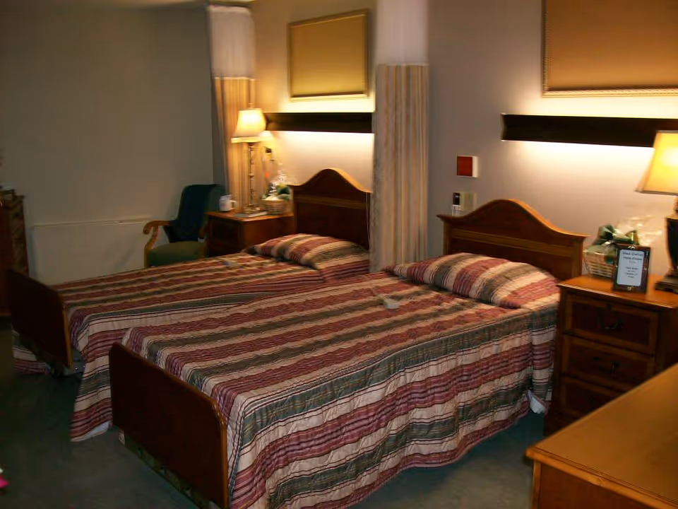Two twin beds with striped bedspreads in a furnished senior living bedroom with nightstands, lamps, and a chair.