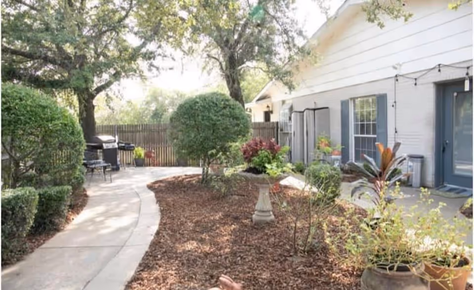 A landscaped outdoor patio area with a curved concrete walkway, trimmed bushes, trees, and various potted plants. There is a white building with blue window shutters and a blue door on the right side. A barbecue grill and outdoor seating are visible near the back fence.