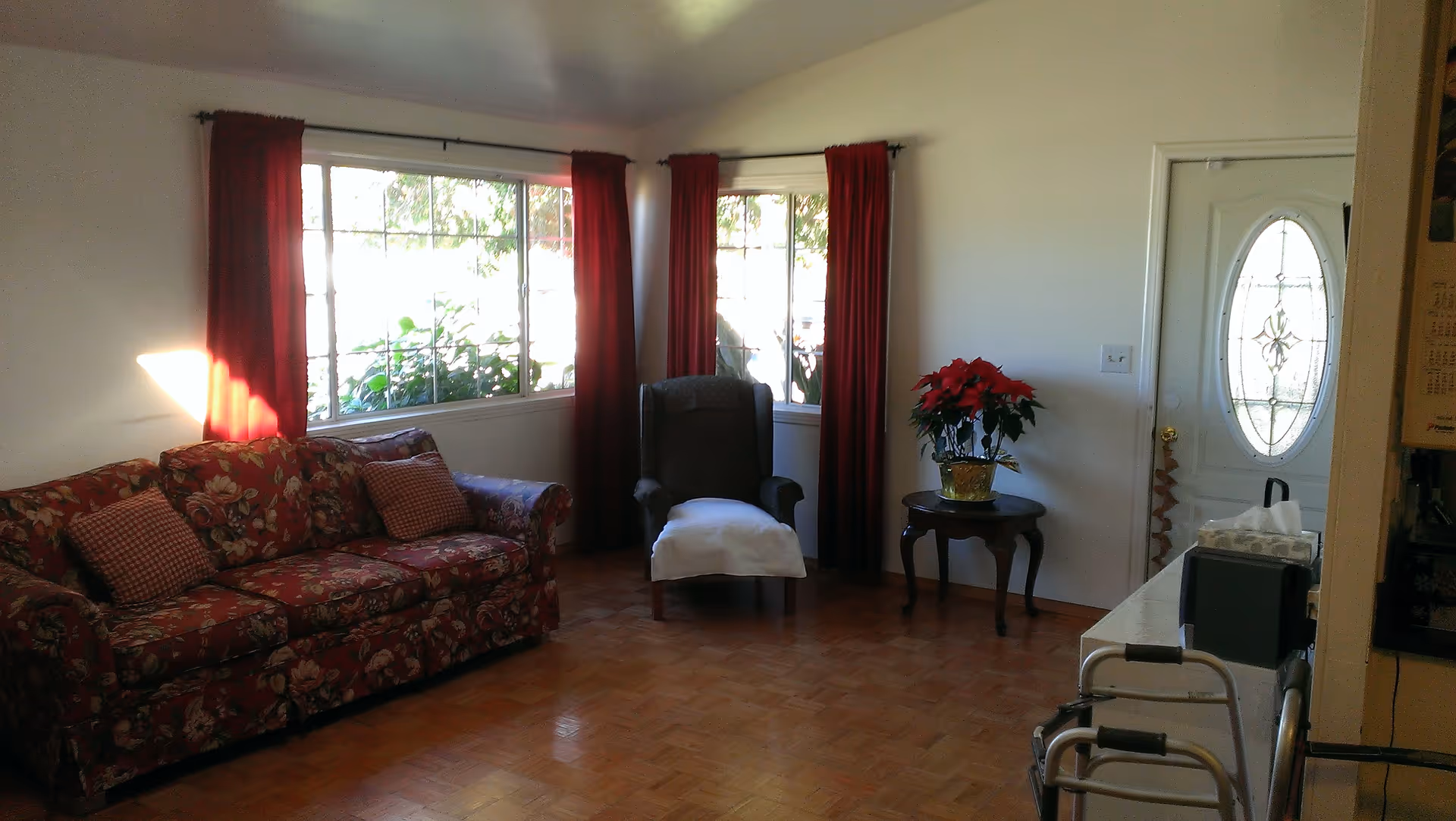 Sunlit living room with a floral-patterned sofa, an armchair by windows with red curtains, a small table holding a poinsettia, and a visible front door.