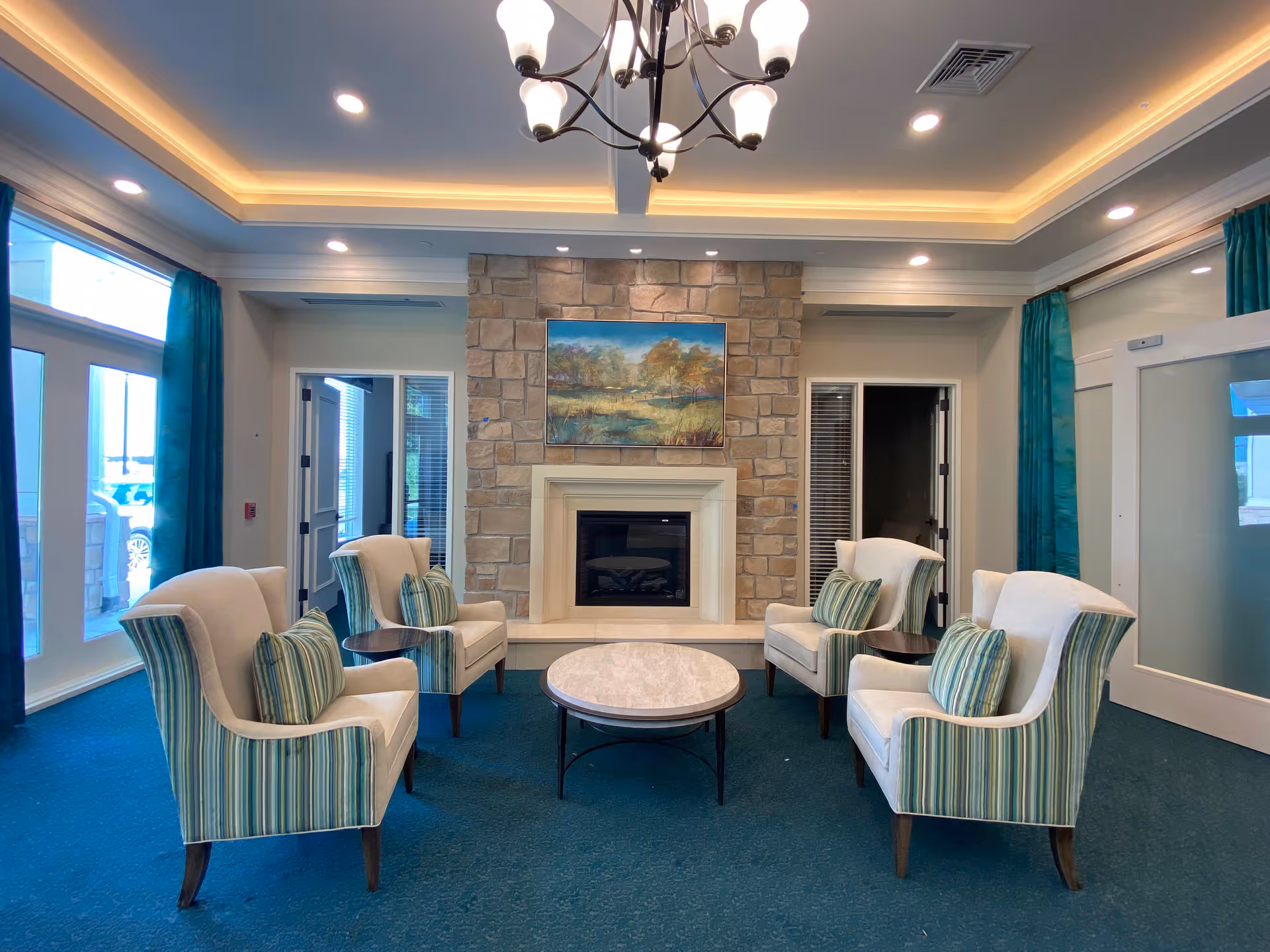 A cozy seating area in a memory care community with four upholstered armchairs arranged around a round coffee table. The chairs have striped fabric on the sides and solid cream-colored backs and seats, each with a striped cushion. Behind the seating area is a stone fireplace with a landscape painting above it. The room has teal carpeting, teal curtains on large windows, recessed lighting, and a chandelier hanging from the ceiling.