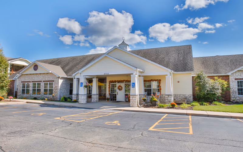 Front exterior view of a single-story senior living facility building with a covered entrance, decorated with fall wreaths and plants. The parking lot in front has designated handicap parking spaces. The sky is blue with some clouds.