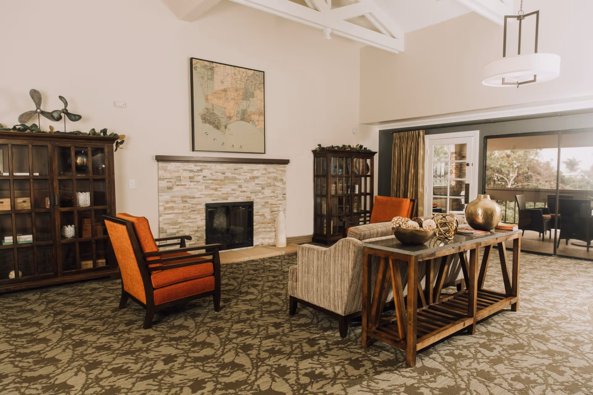 A cozy living room with a patterned carpet, two orange armchairs, a striped armchair, and a wooden console table with decorative items. There is a stone fireplace with a framed map above it, two glass-front cabinets on either side, and large sliding glass doors leading to an outdoor patio.