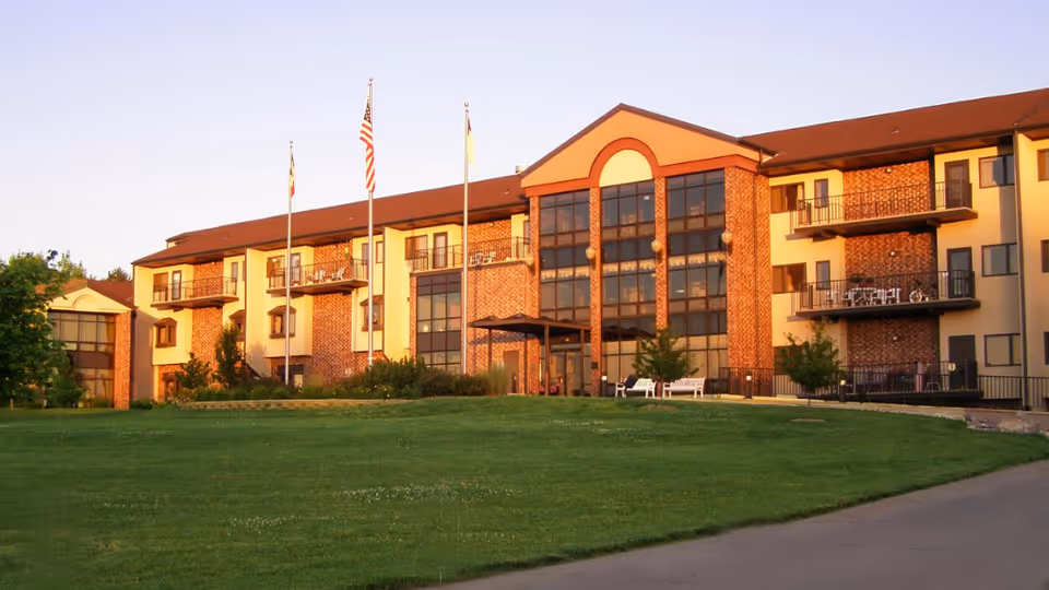 Front exterior of a multi-story senior living building with balconies, flagpoles, and a large grassy lawn.